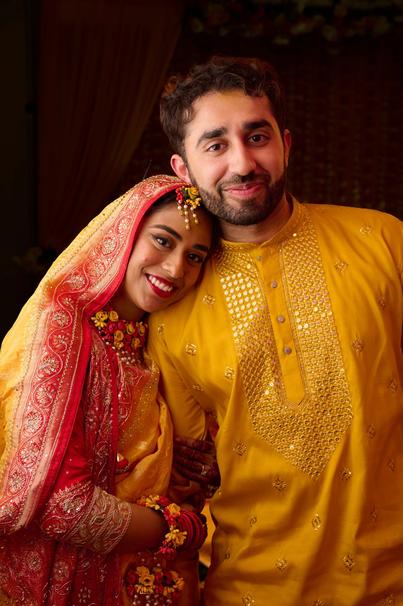 Bride in yellow and red dupatta leaning on groom in yellow kurta, both smiling warmly